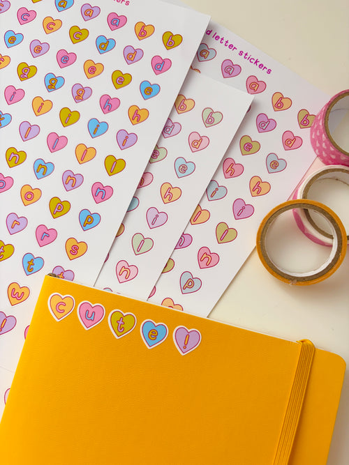 Colorful heart-shaped stickers on a white sheet with a yellow notebook and tape in the foreground.