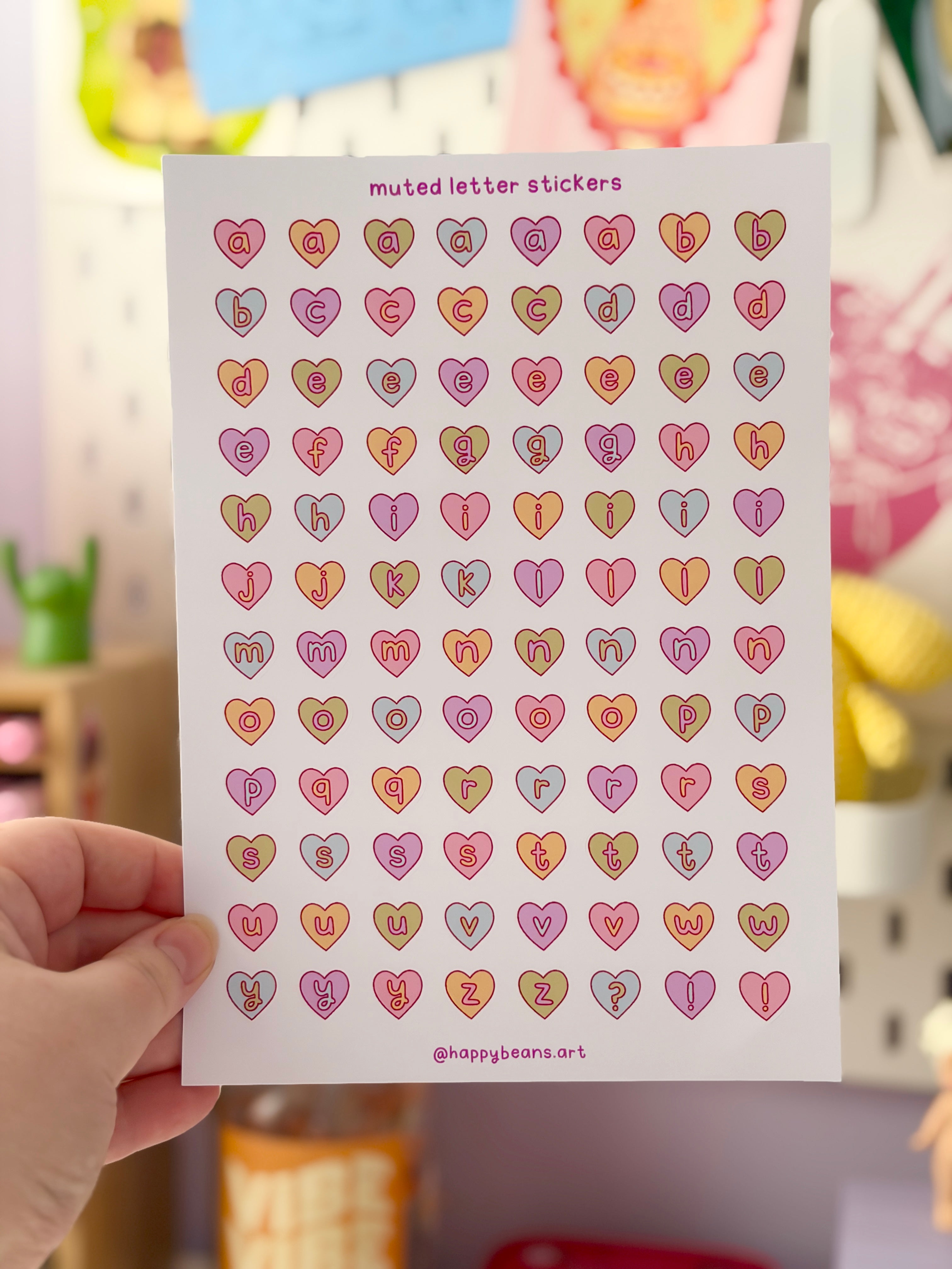 Sheet of heart-shaped stickers with letters held by a hand in a blurred indoor setting.